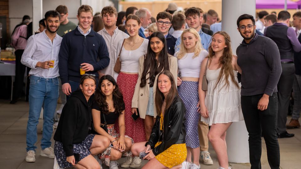 Photo of a group of students looking at the camera smiling, stood outside the Edward Herbert Building on the University campus. Some are standing, and some are kneeling on the floor