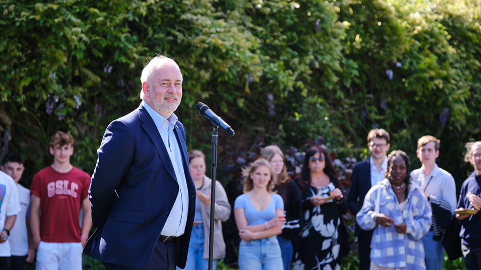 Photo of Vice-Chancellor Professor Nick Jennings stood on an outside stage in front of a microphone smiling with students in the background watching 