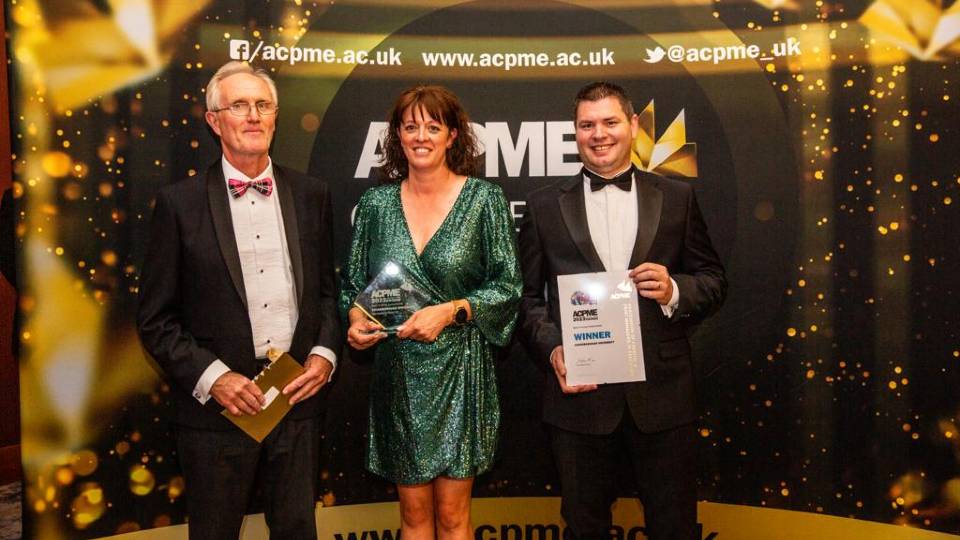 Photo of Helen Clarke wearing a green dress and Paul Goddard, Assistant Manager in Print Post and Logistic Services wearing a black suit. Both are smiling at the camera holding a framed award they have just won