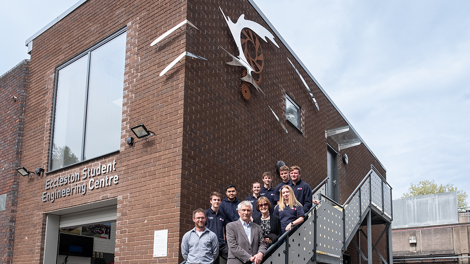Photo of alumnus Ian Tricker, Barry and Valerie Eccleston and students who form part of the LU Motorsport team, standing on stairs of the Eccleston Building with Ian Tricker's sculpture above them on the exterior wall