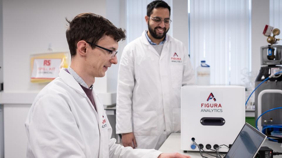 Two people wearing lab coats working on a laptop in a lab.