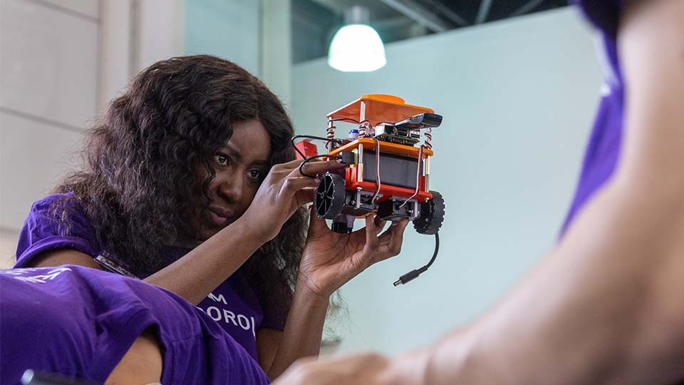 Photo of a woman wearing a 'Team Loughborough' purple tshirt staring in concentration as she looks at a car-type object she is holding in her hands