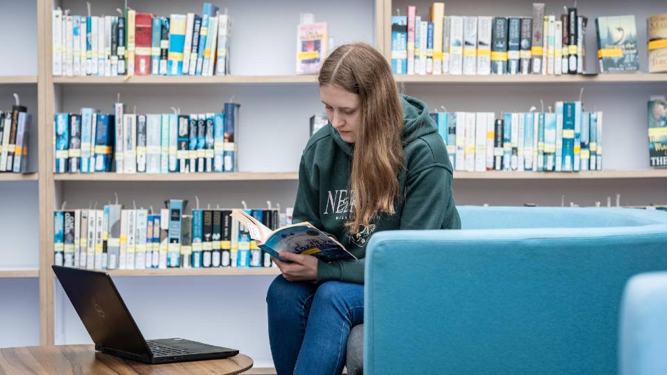 A postgraduate student sitting and reading a book in the Library with their laptop on the table in front of them.