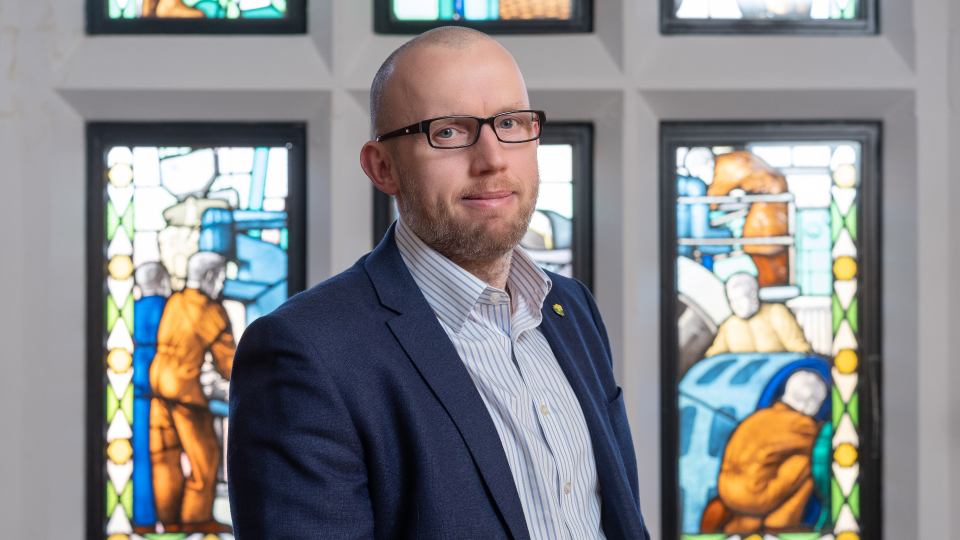 Photo of Martyn Edwards stood on his side with his face looking towards the camera. He is wearing a dark suit and is stood in front of stained glass windows inside the Hazlerigg Building