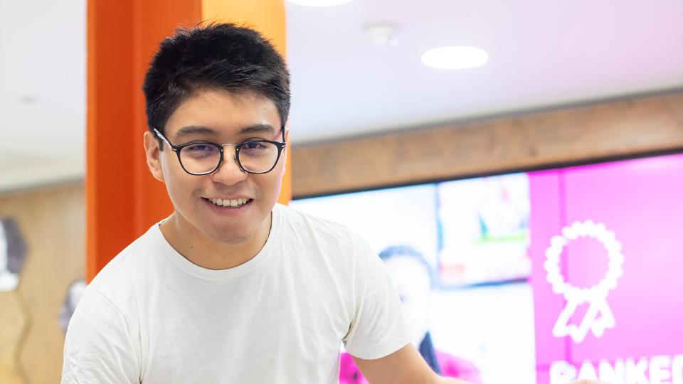 A male student wearing glasses and a white t-shirt is stood over a table and behind a large digital screen smiling towards the camera