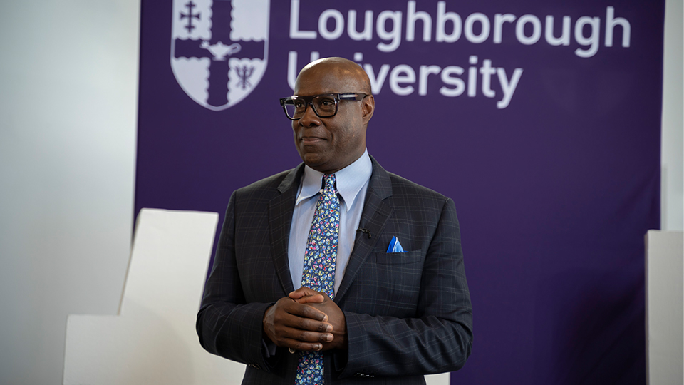 Photo of Mike Wedderburn standing up wearing a suit at the Loughborough campus, in front of a sign showing the University logo