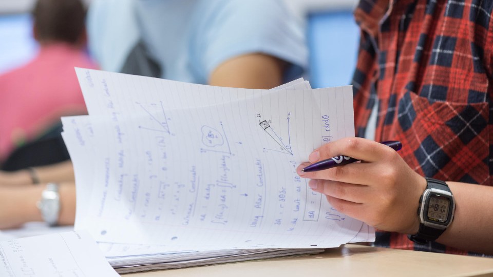 Person holding a pen and a stack of papers with handwritten mathematical equations, diagrams, and formulas.