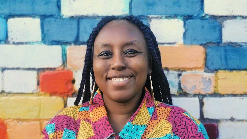 Nadina Ali smiling in front of a wall of white, blue, red and yellow painted bricks.