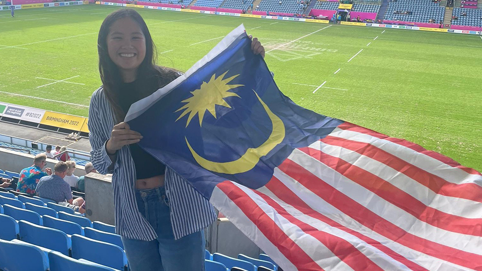 Naomi Tan holding a flag in stands at Rugby 7's stadium at the Commonwealth Games