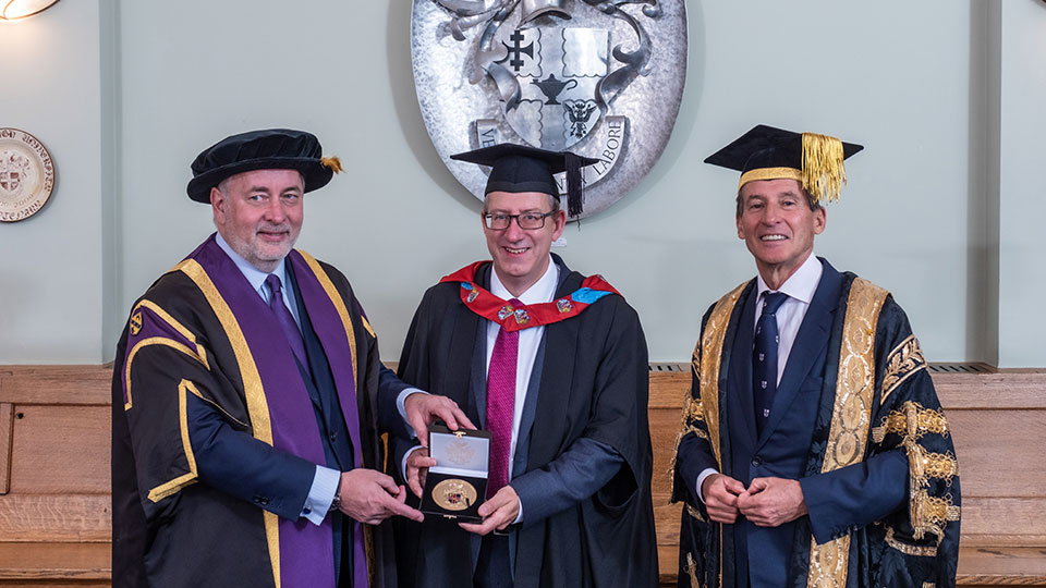 Three men in academic robes with the man in the middle holding a medal