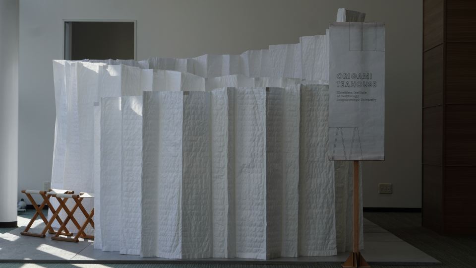photo of the mobile origami teahouse designed by Loughborough students. The teahouse is made from white material, and has a banner in front of it, along with stools inside. 