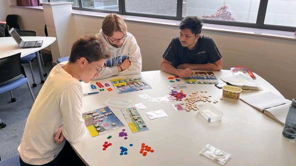 photo of three male students sat at a table working on a puzzle that has different coloured counters and various materials 