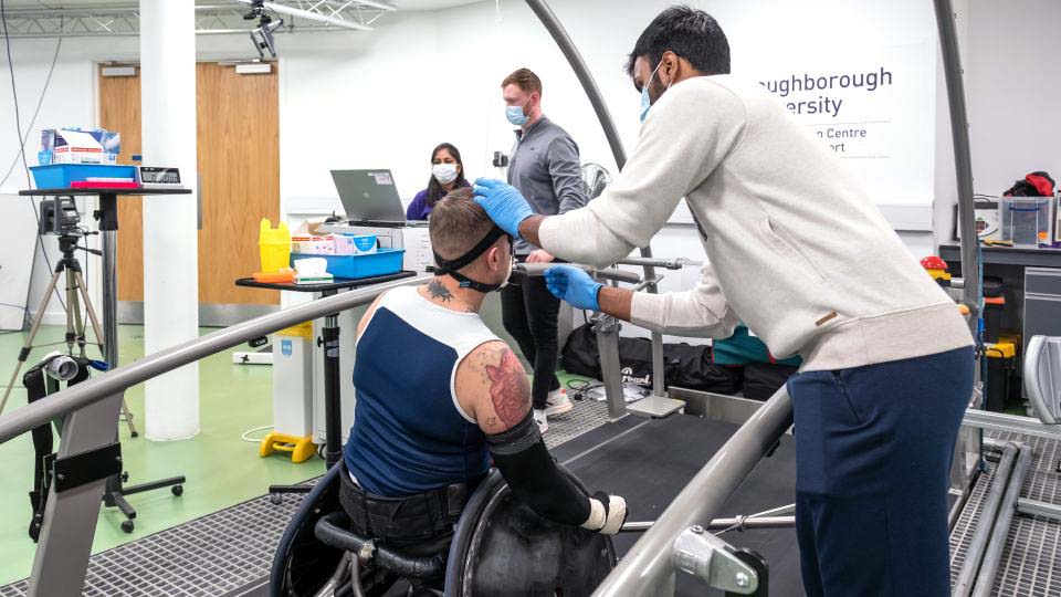 Photo of a researcher wearing PPE whilst testing a man sat in a wheelchair on top of some equipment in a lab environment with other researchers looking at a laptop in the background