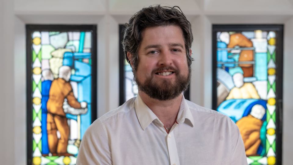 Richard smiling at the camera wearing a white shirt, sat inside the Hazlerigg Building with stained glass windows behind him
