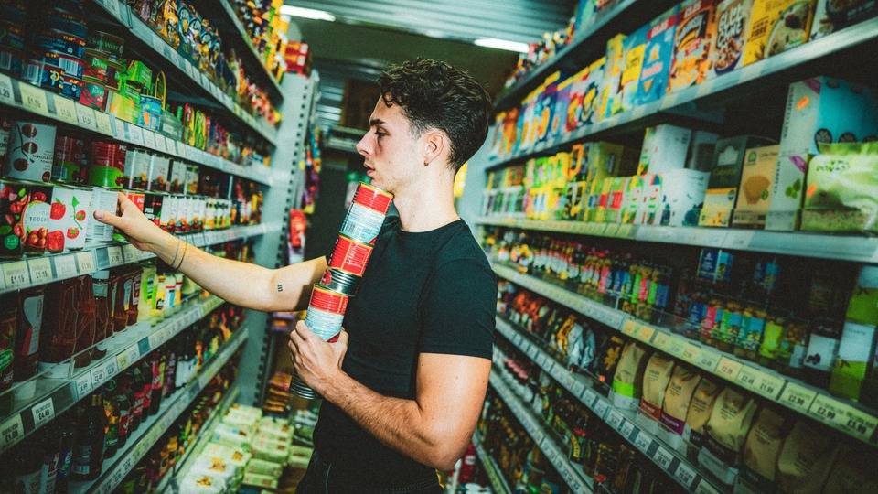 A student holding a stack of tins in the aisle of a supermarket.