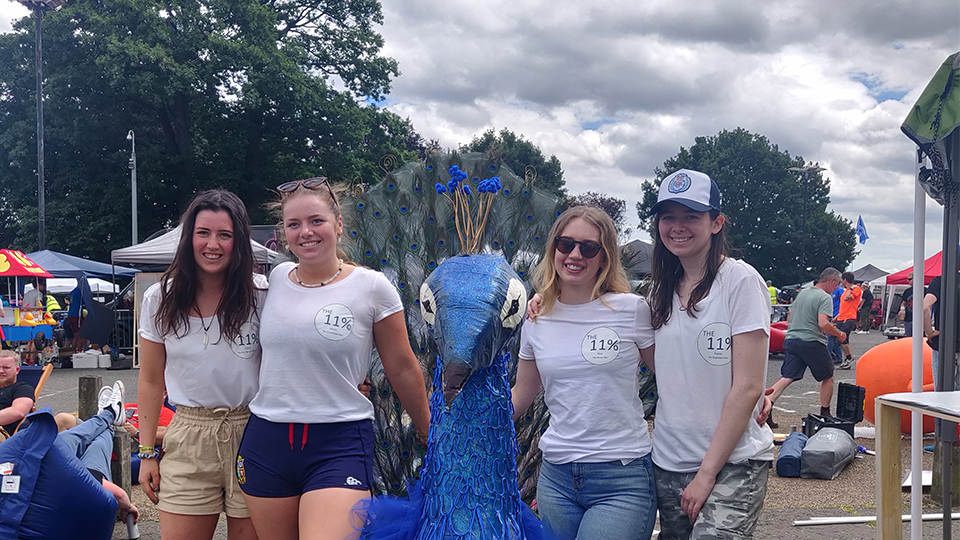 Four students standing behind their soapbox - a blue peacock with wheels.