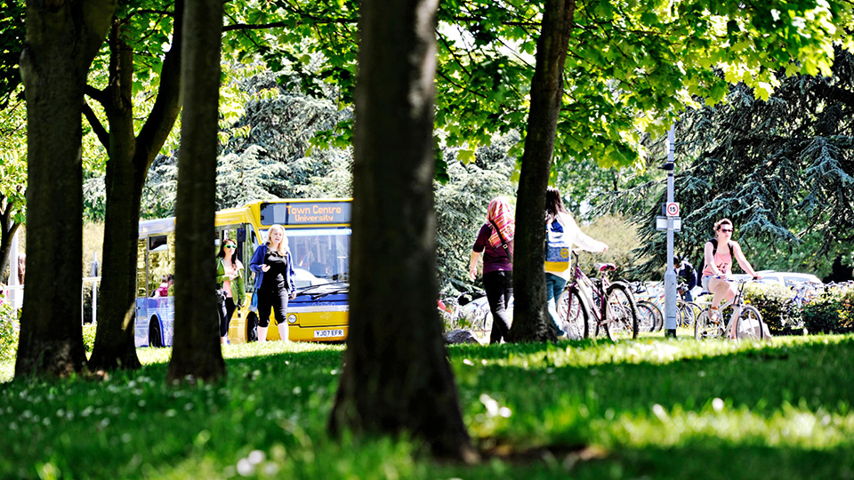 A photo of the Sprint yellow and blue bus driving up a road on campus with lots of trees around and students walking and cycling on bikes 