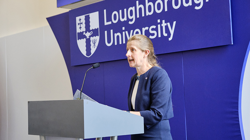 Side profile of Professor Rachel Thomson speaking into a microphone on a stand at an event, with the Loughborough University logo behind her
