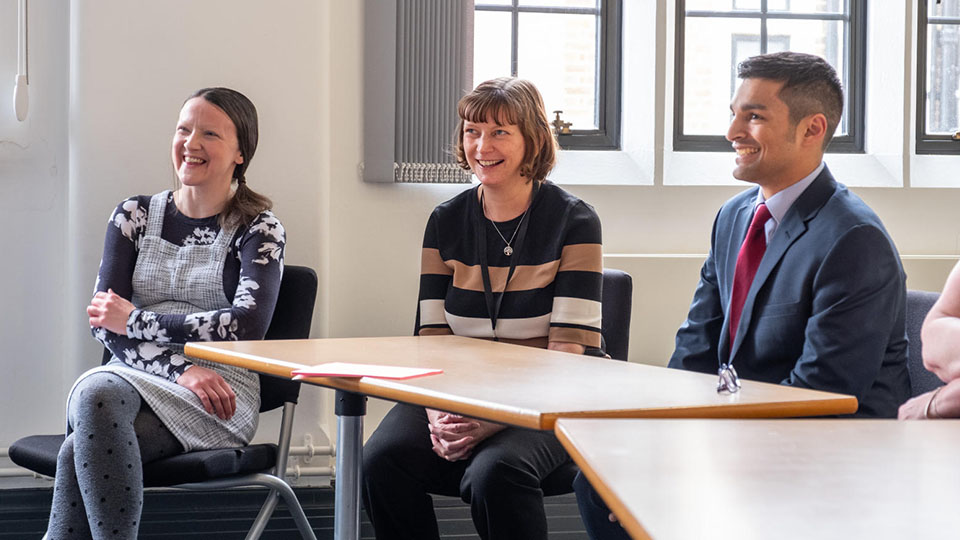 Photo of three members of staff sat down by tables smiling whilst watching a presentation