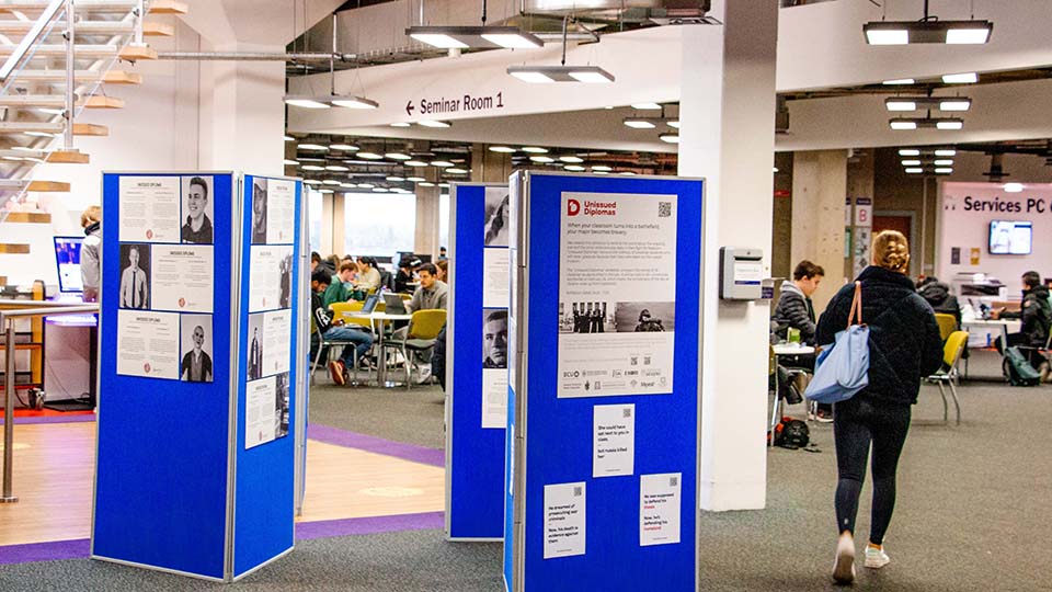 Photo of the Unissued Diplomas exhibition on blue boards inside the University Library, with a student walking past