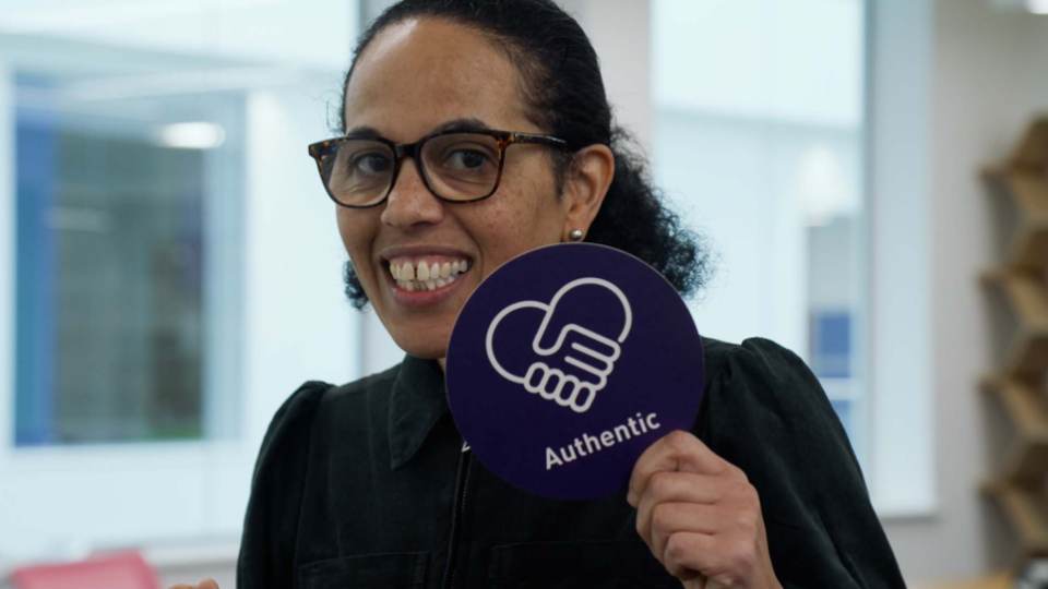 A staff member smiling at the camera holding a round, purple sign that says 'Authentic' with a icon of two hands holding in the shape of a heart