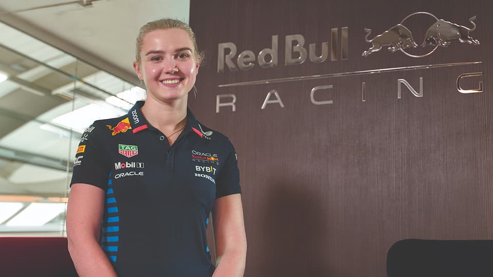 A smiling woman stands in front of a dark wood wall featuring the silver Red Bull Racing logo. She is wearing a navy blue team polo shirt with various sponsor logos.