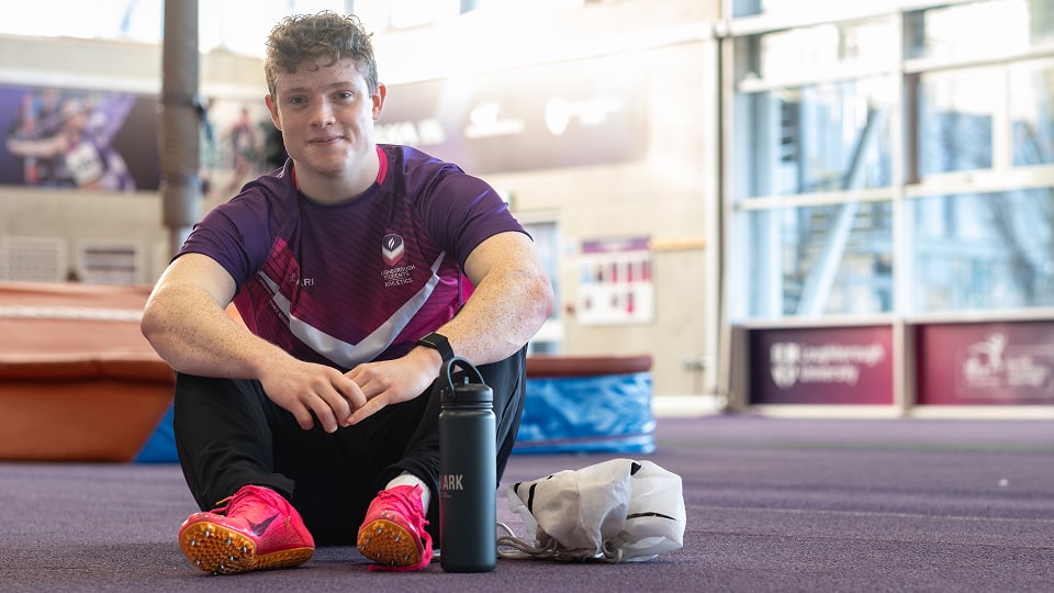 An athlete in a purple track shirt and bright pink sprinting spikes sits on an indoor track. He is resting with a water bottle and gym bag next to him, with high-jump mats and large windows visible in the background.