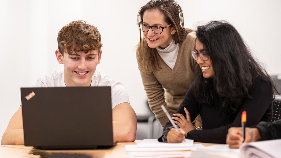 A teacher wearing glasses leans in to look at a laptop screen with two smiling students. They are sitting at a desk in a bright classroom setting.