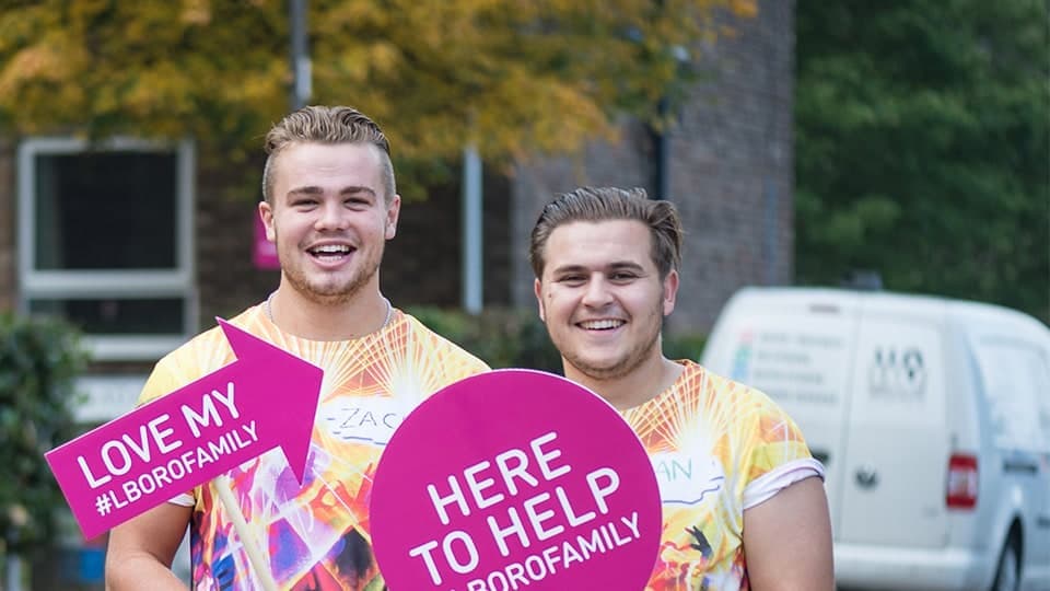 Two people are smiling and standing in front of a building holding pink signs, providing support for students moving in